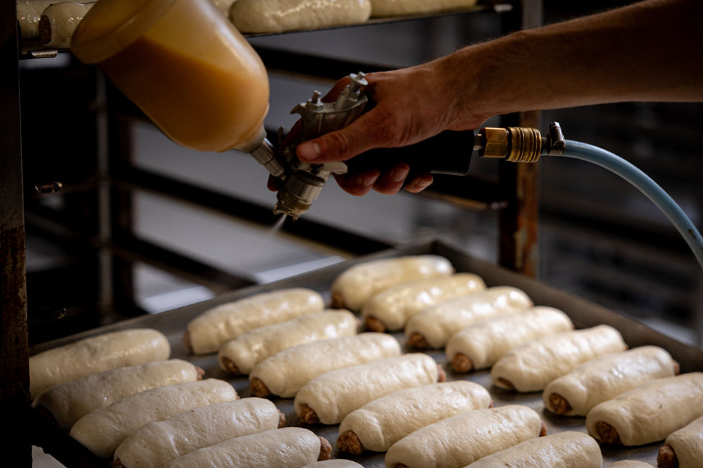 Bakker spuit rijen worstenbroodjes af met een laagje ei voor de glans